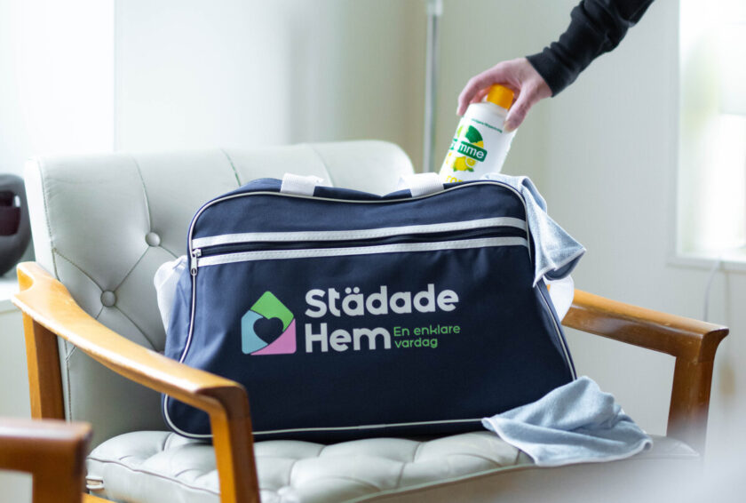Städväska med texten "Städade Hem" på en fåtölj, hand lägger rengöringsmedel ovanpå.